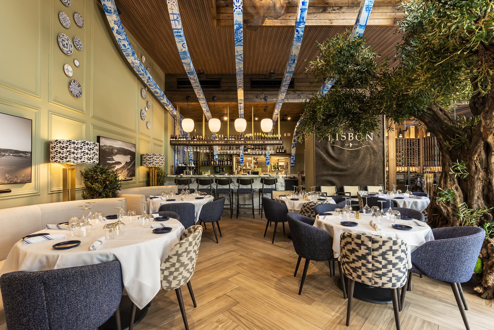 Old Lisbon dining room: white-clothed tables, navy and patterned chairs, blue azulejo-tile ceiling beams, an olive tree and the bar with the Old Lisbon sign behind