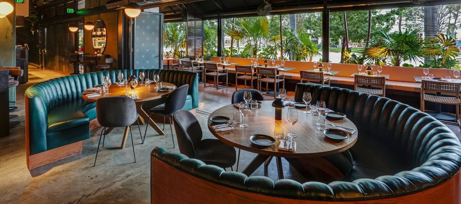 Orilla's dining room: curved green leather booths around round wooden tables set with glassware, with floor-to-ceiling windows onto tropical palms